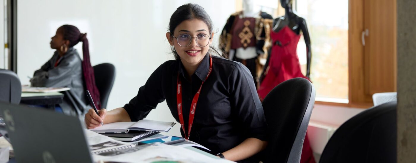A student sits at a table in a fashion studio in the Creative Hub. She is holding a pen and writing in a notebook. There is an open laptop on the table in front, and two dressed mannequins behind.