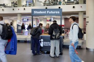 Two students talk to a staff member at a black 'Student Futures' stand. The stand is on the gorund floor of the Learning Hub and other students walk past.