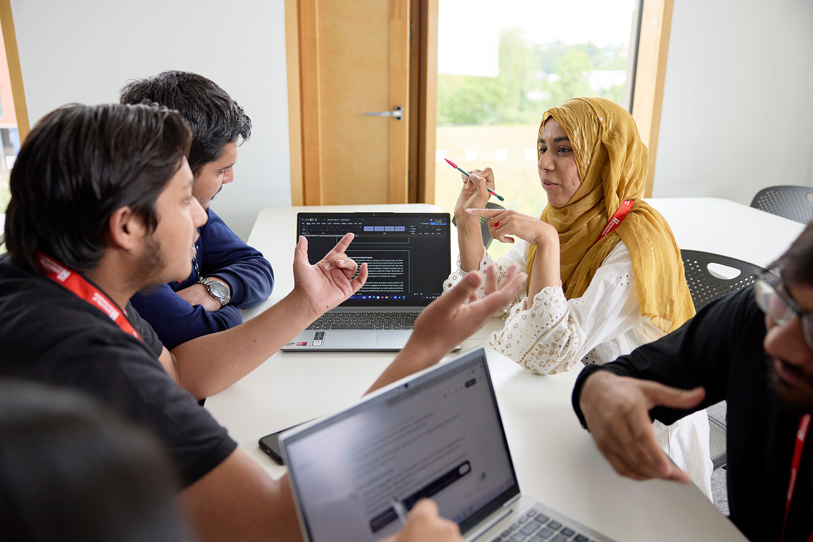 Four students taking part in a discussion at a table, with a couple of laptops in front of them. Their hands are raised while they talk through their points.