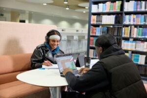 Image of two students sat opposite one another in a library booth chatting, headphones on the head of the student facing the camera.
