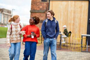 An image of three students walking along outside the engine shed, the student in the middle is holding a coffee cup