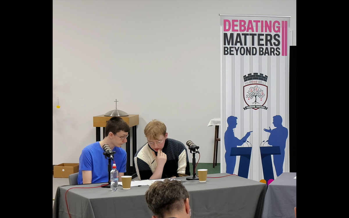 Two participants in the UON AI debate held at HMP Five Well prison sit at a table and look at their notes. On the tabelk are two microphones, coffee cups, and shhets of paper. Behind them is a pull-up banner which reads 'Debating Matters Beyond Bars'.