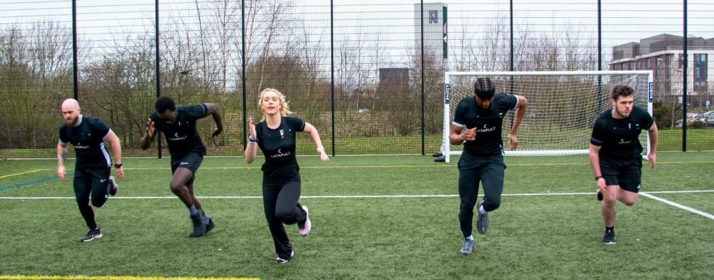 Five students sprinting forward on an outdoor artificial grass pitch, running toward the camera from a starting position, with a football goal, tall perimeter fencing, and campus buildings visible in the background.