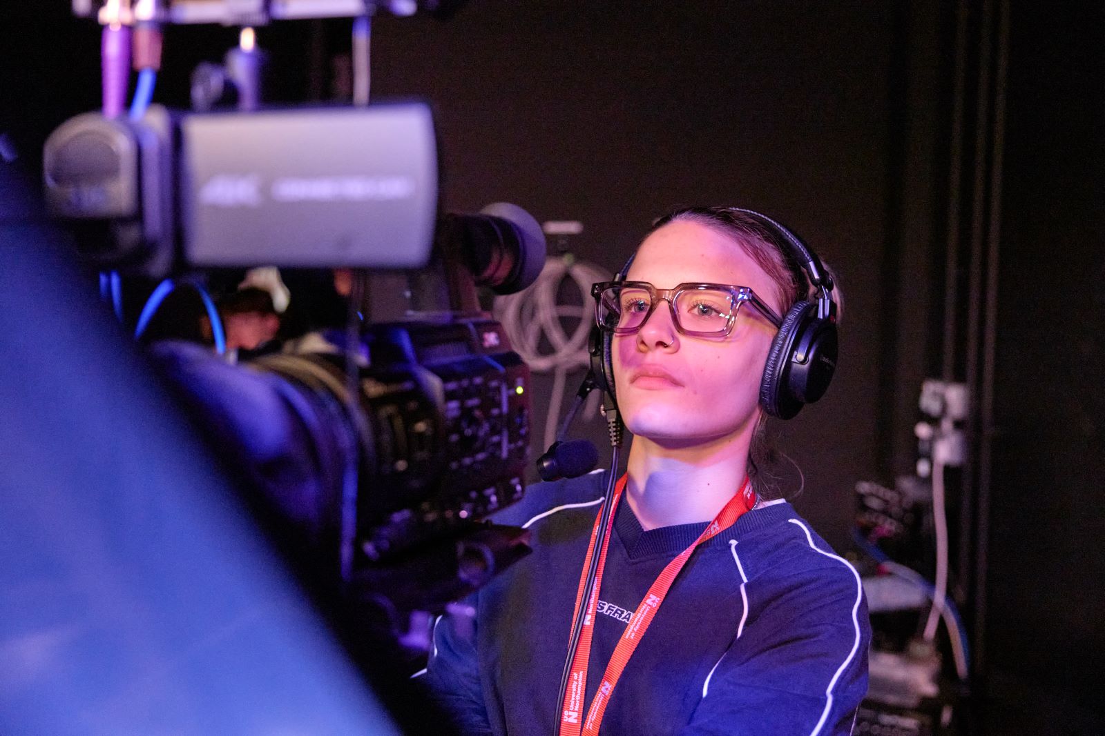 Student wearing glasses and a headset operates a professional video camera in a dimly lit studio with coloured lighting and production equipment.