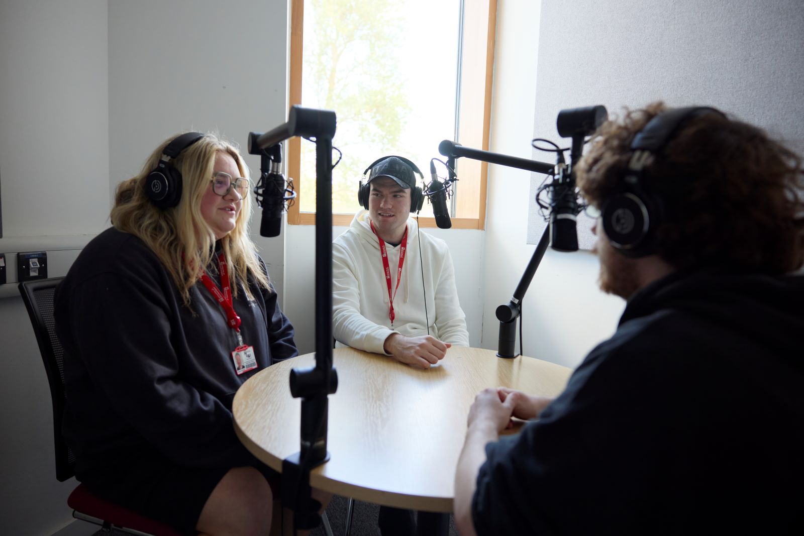 Three students record a podcast next to a window in a small room. They wear headphones and are sitting around a table in front of microphones.