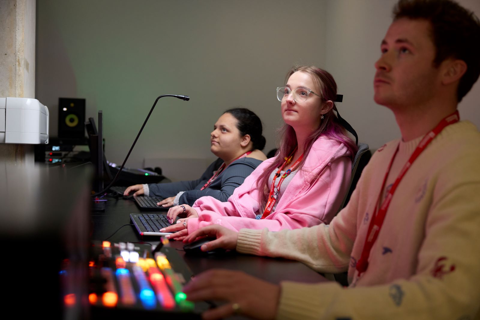 Three students wearing red lanyards work at a production desk, using keyboards and a mixing console in a TV studio control room.