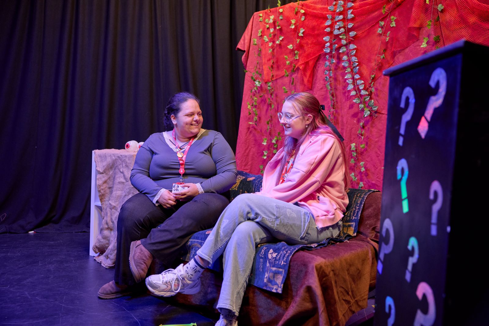 Two students sit on a sofa on the set of a TV studio. They both wear red lanyards. The sofa and the background are draped with coloured cloth.