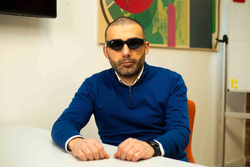 Man wearing tinted glasses is sat at a desk, looking at the camera.