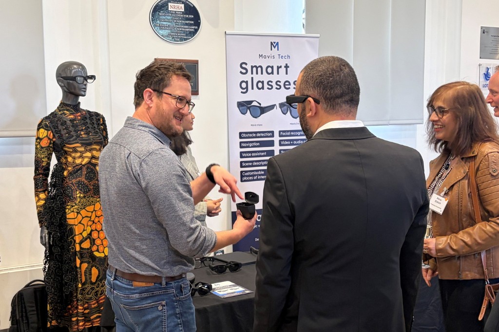 Two people have a conversation while holding a pair of tinted glasses. One smiles at the other, and there is a banner which says 'smart glasses' behind them.
