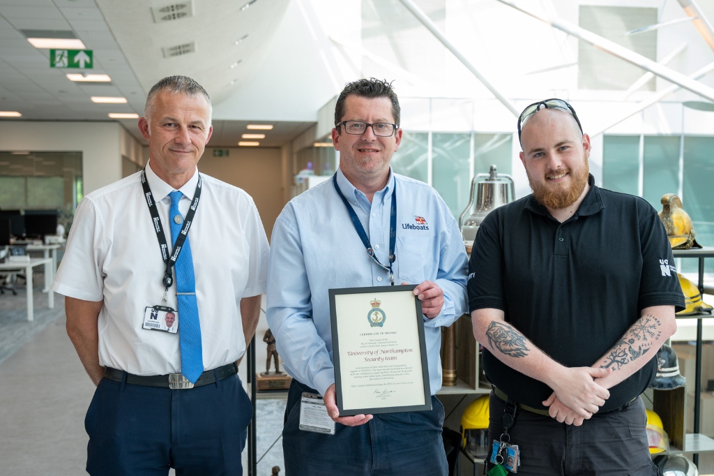 Three people stand next to each other and smile at the camera. The person in the centre holds a certificate. The background is a light office building.