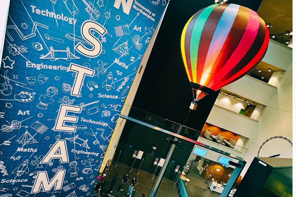 An indoor remote-controlled model hot air balloon fires its burner as it passes a sign on the third floor of the University of Northampton's Learning Hub displaying the word STEAM.