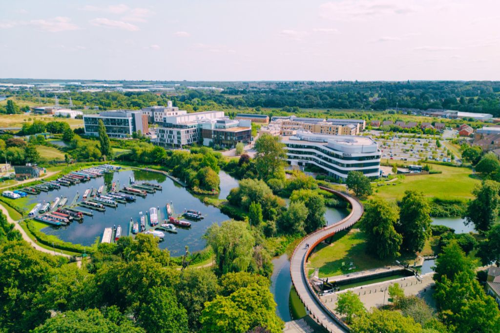 Aerial image of UON's Waterside Campus with a canal boat marina in the foreground disected by the bridge snaking its way from Beckets Park to the modern buildings of Waterside, the background filled with the leafy hinterlands south of Northampton.