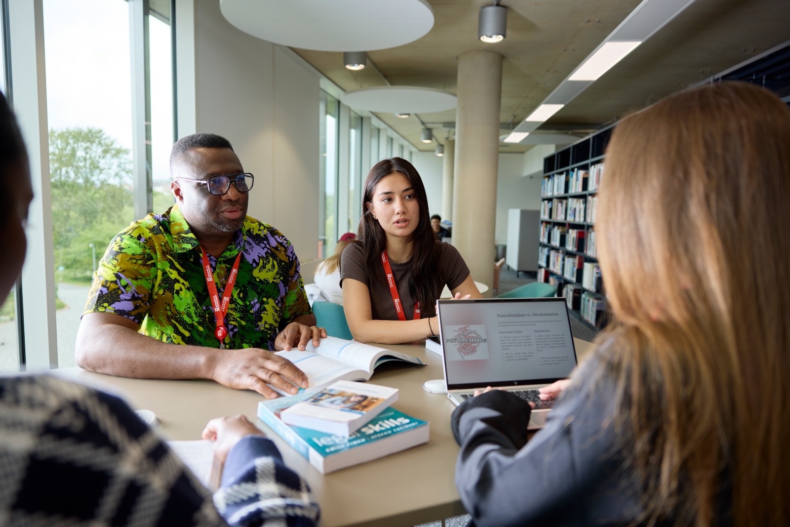 Students studying together around a table in the University of Northampton library, discussing coursework with books and a laptop.