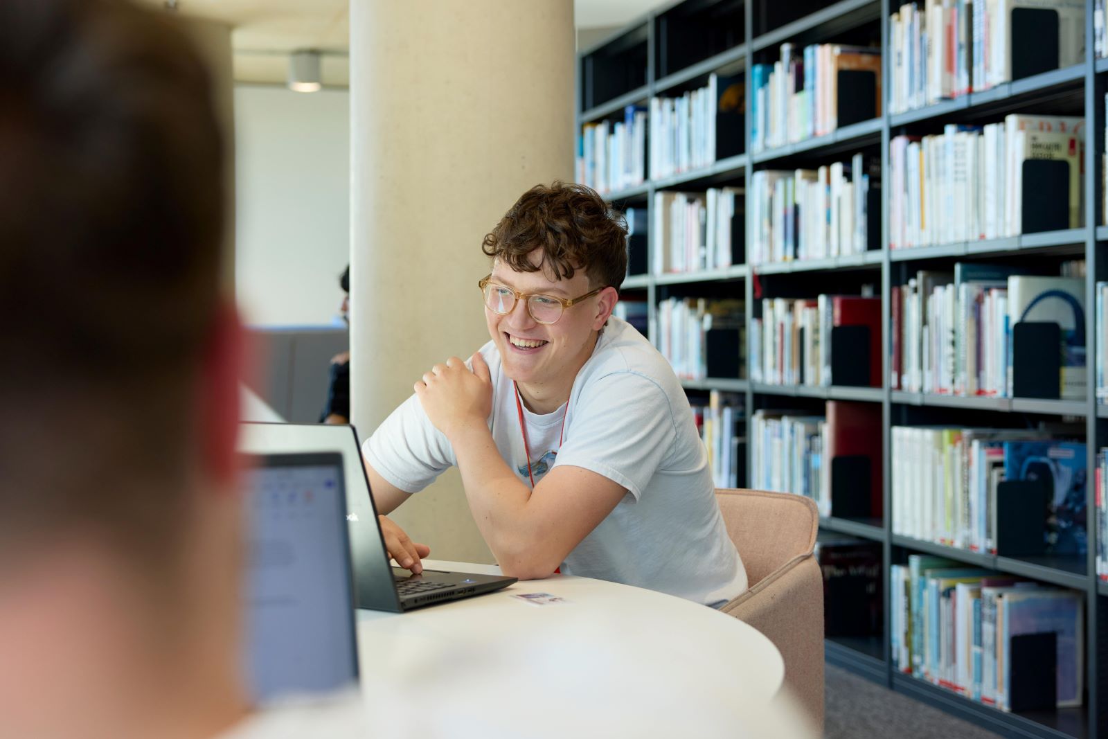 A student works on a laptop in front of a bookstack. He is wearing a t-shirt and is smiling. In the foreground is the blurred head of another student seated at the same table.
