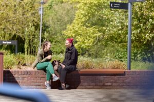Two female students sit on an outside wall on Waterside Campus on a sunny day. They are talking and laughing.
