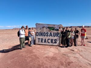 A group of students pose for a photo by a large 'Dinosaur Tracks' sign. It is a sunny day. The students are casually dressed in t-shirts, shorts and sunglasses. They are surrounded by desert rocks.