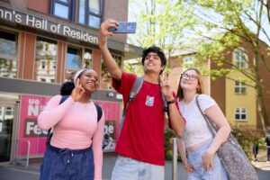 Three students pose for a selfie in front of St Johns Halls. It is a sunny day, and all three are wearing t-shirts.