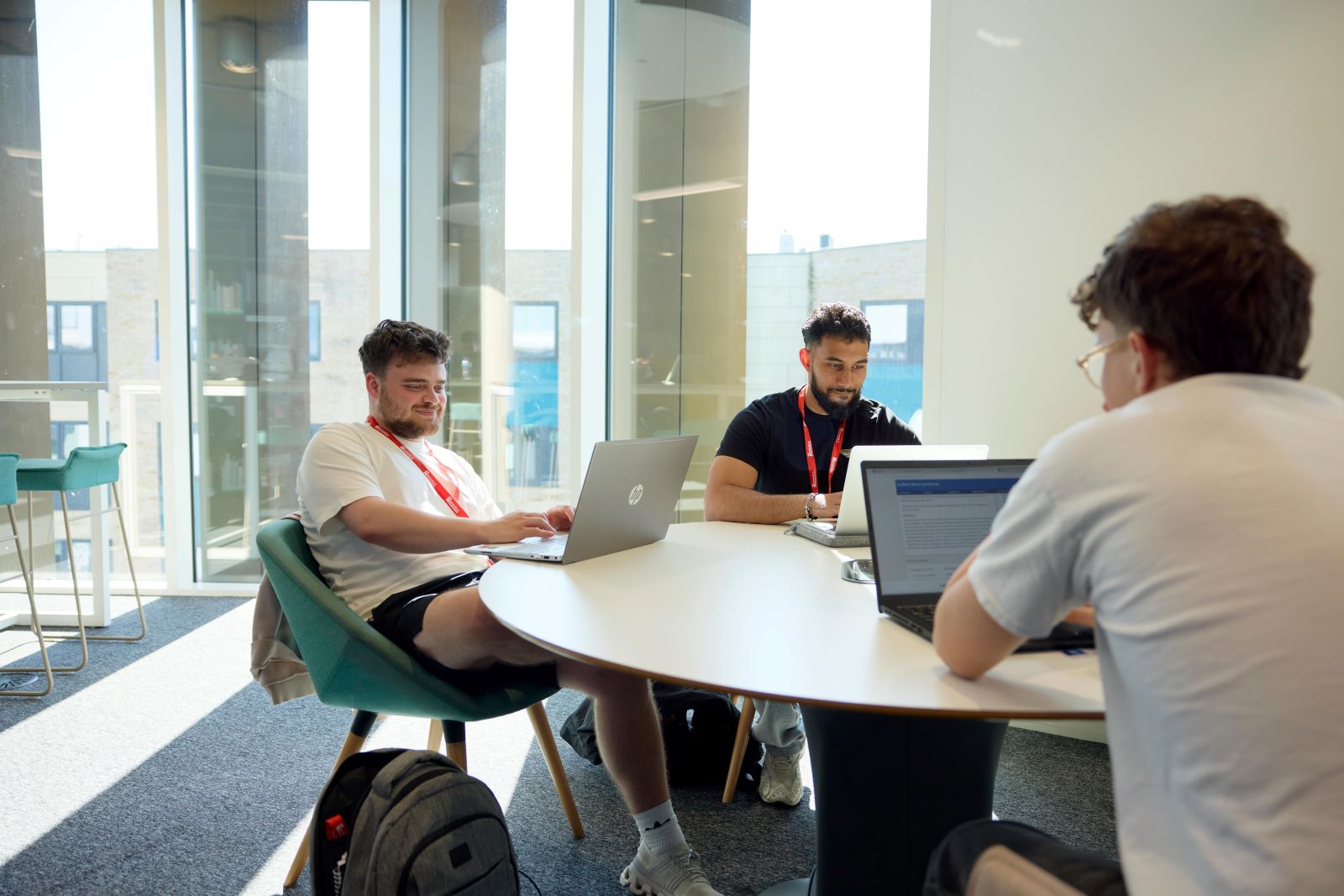 Three students work on laptops on a round table in the Learning Hub. They are casually dressed in t-shirts. Behind them are large floor-to-ceiling windows.