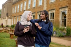 Two students stand together in a garden next to a large stone building. One is wearing a hijab and a cardigan and holds a travel mug; the other is wearing a sweatshirt and is resting her arm on the shoulder of her friend. They are both carrying rucksacks and are smiling.