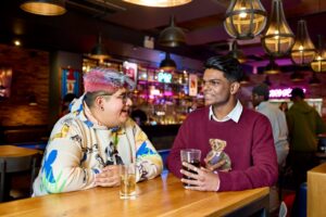 Two students sit at a table with drinks at Waterside bar and restaurant. Behind them is the bar, which is blurred.