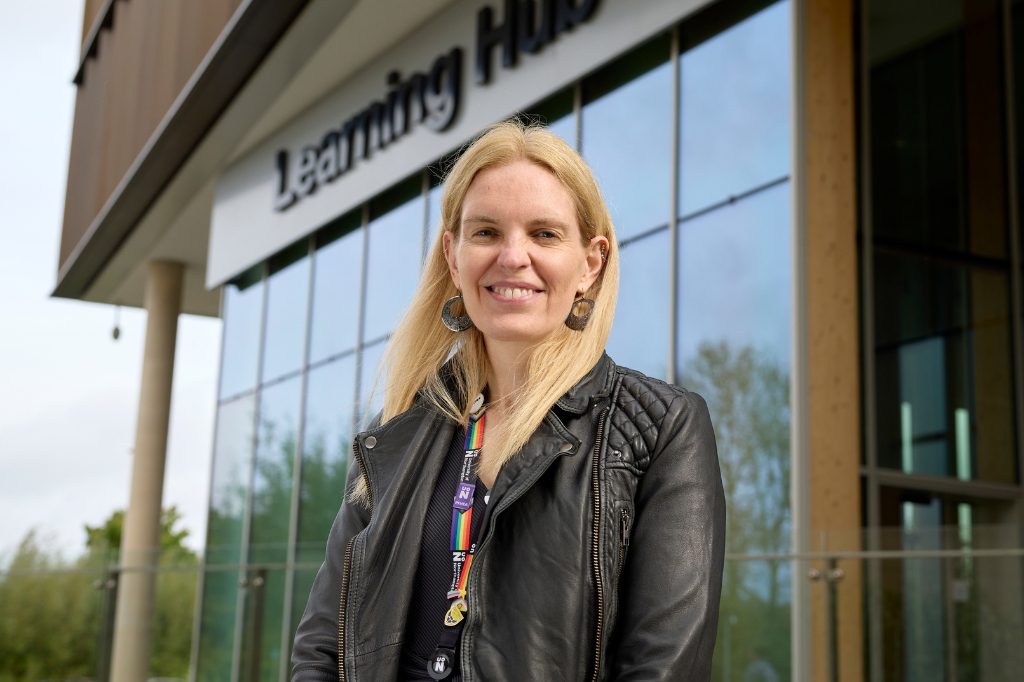 Person smiles at the camera outdoors in front of a glassed building.