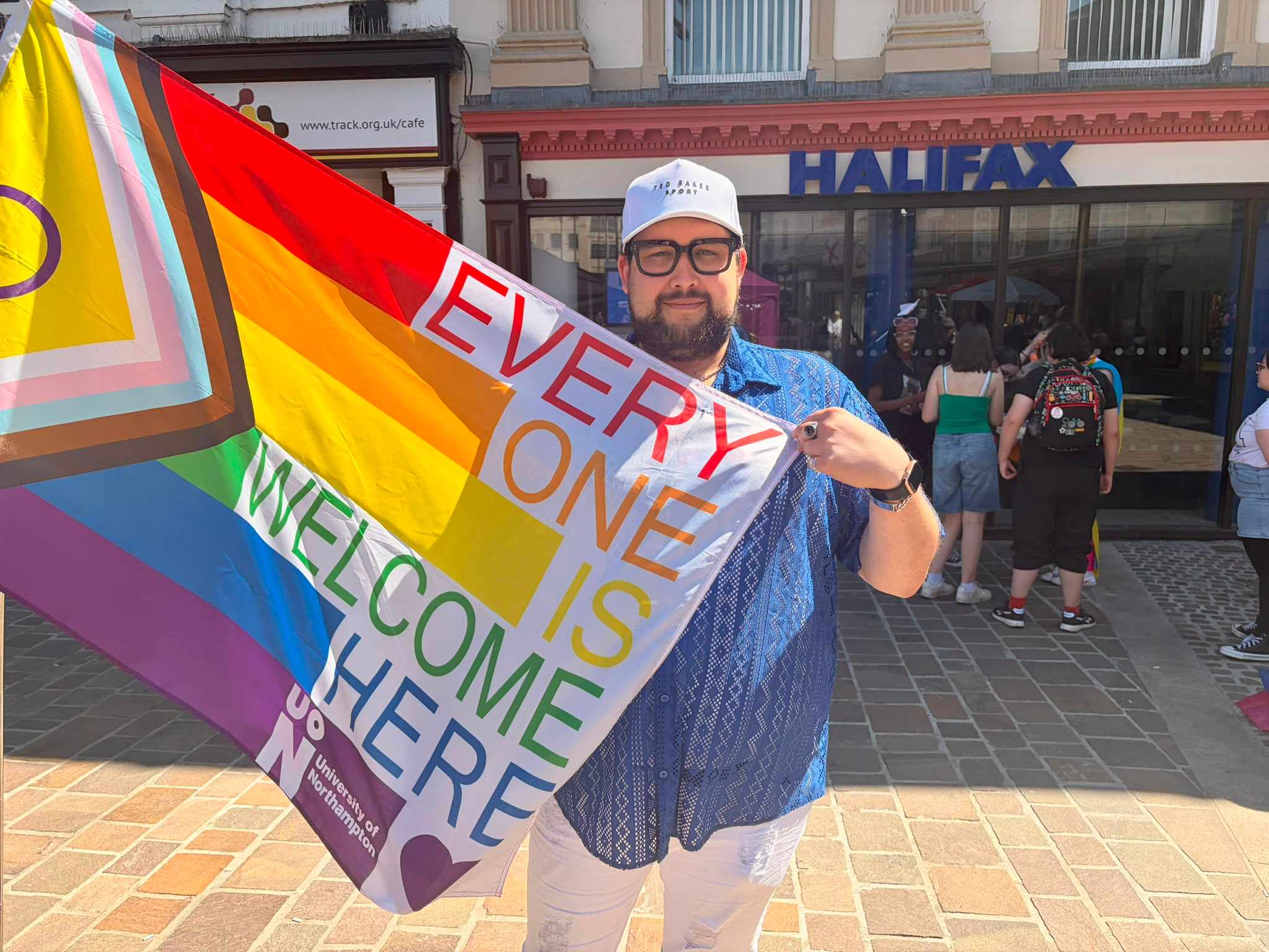 Anthony stands in front of Halifax building, smiling at the camera and holding aflag which reads 'every body is welcome here'