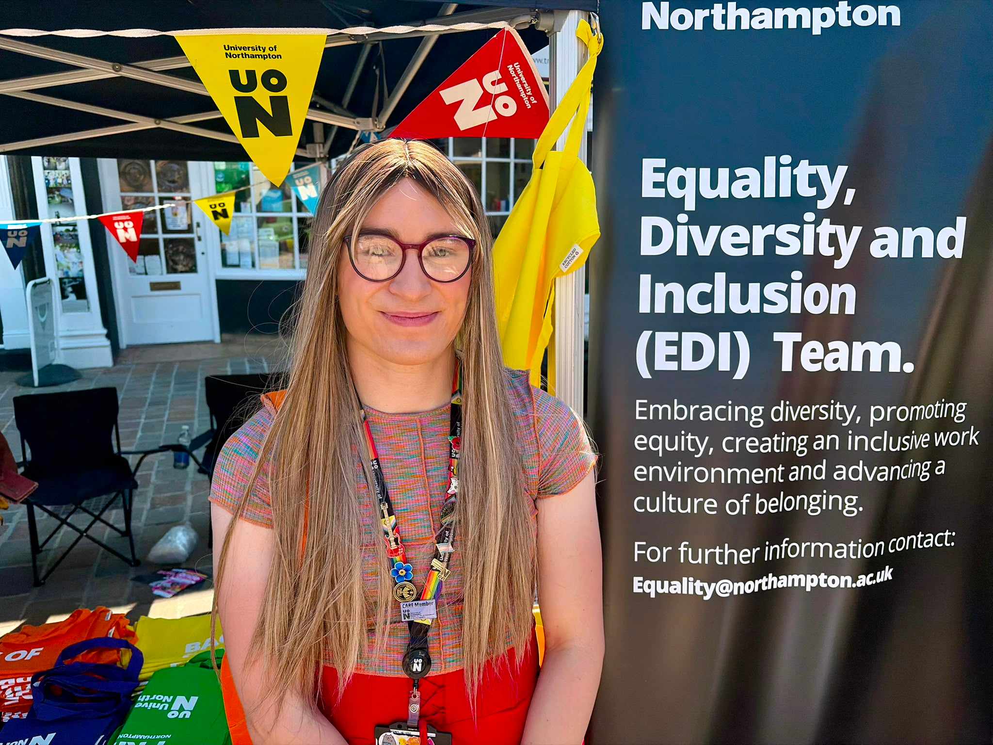 Sophie-Mai smiles to camera in front of a black 'University of Northampton' gazebo, standing next to a sign which reads 'Equality, Diversity and Inclusion (EDI) team'