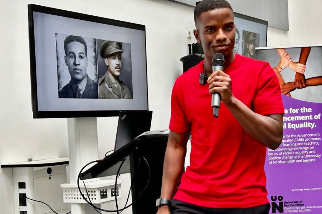 Leadership and personal development specialist Ollie Wanjohi stands imposingly with a mircophone as he talks about the life of Walter Tull, images of whom are displayed on screens behind Ollie.
