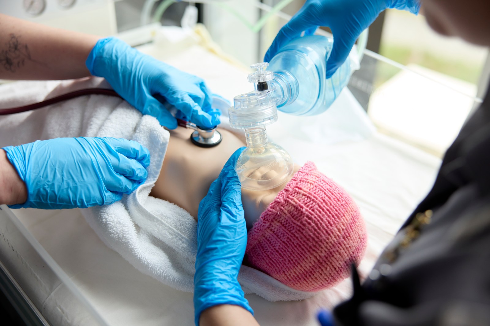 Close-up of gloved hands caring for a newborn, using a stethoscope and resuscitation mask during a neonatal training procedure.