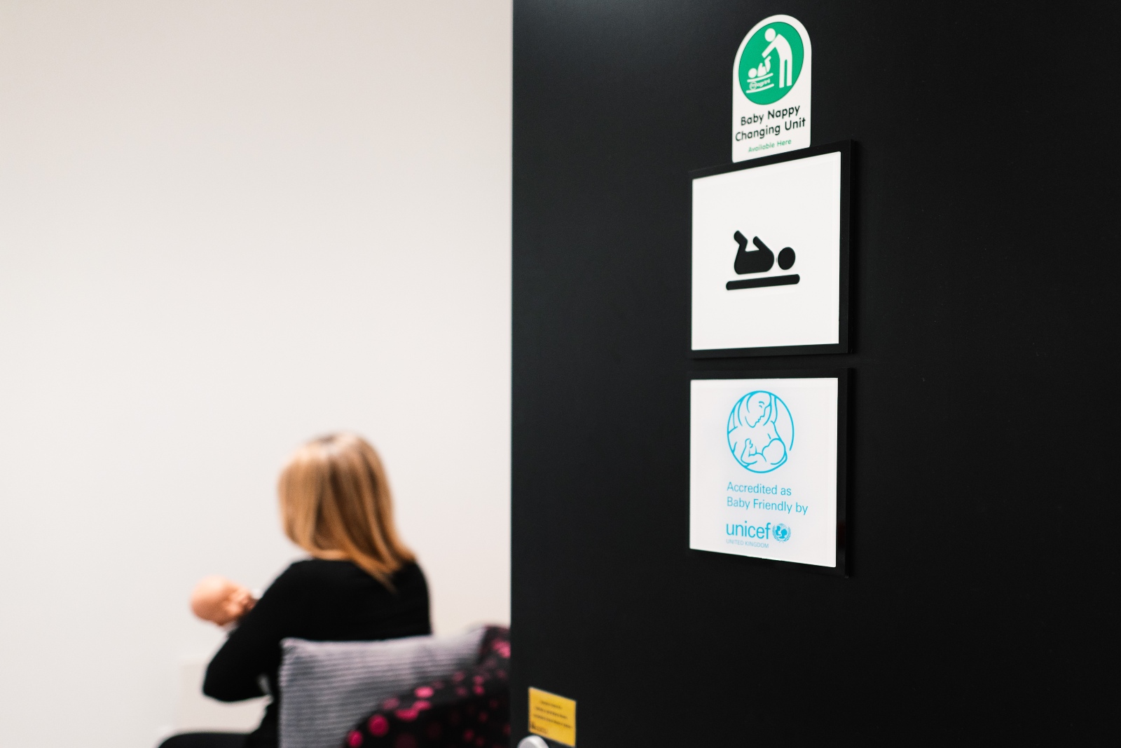Signage for baby changing facilities and Baby Friendly accreditation beside a room where an adult is holding an infant.