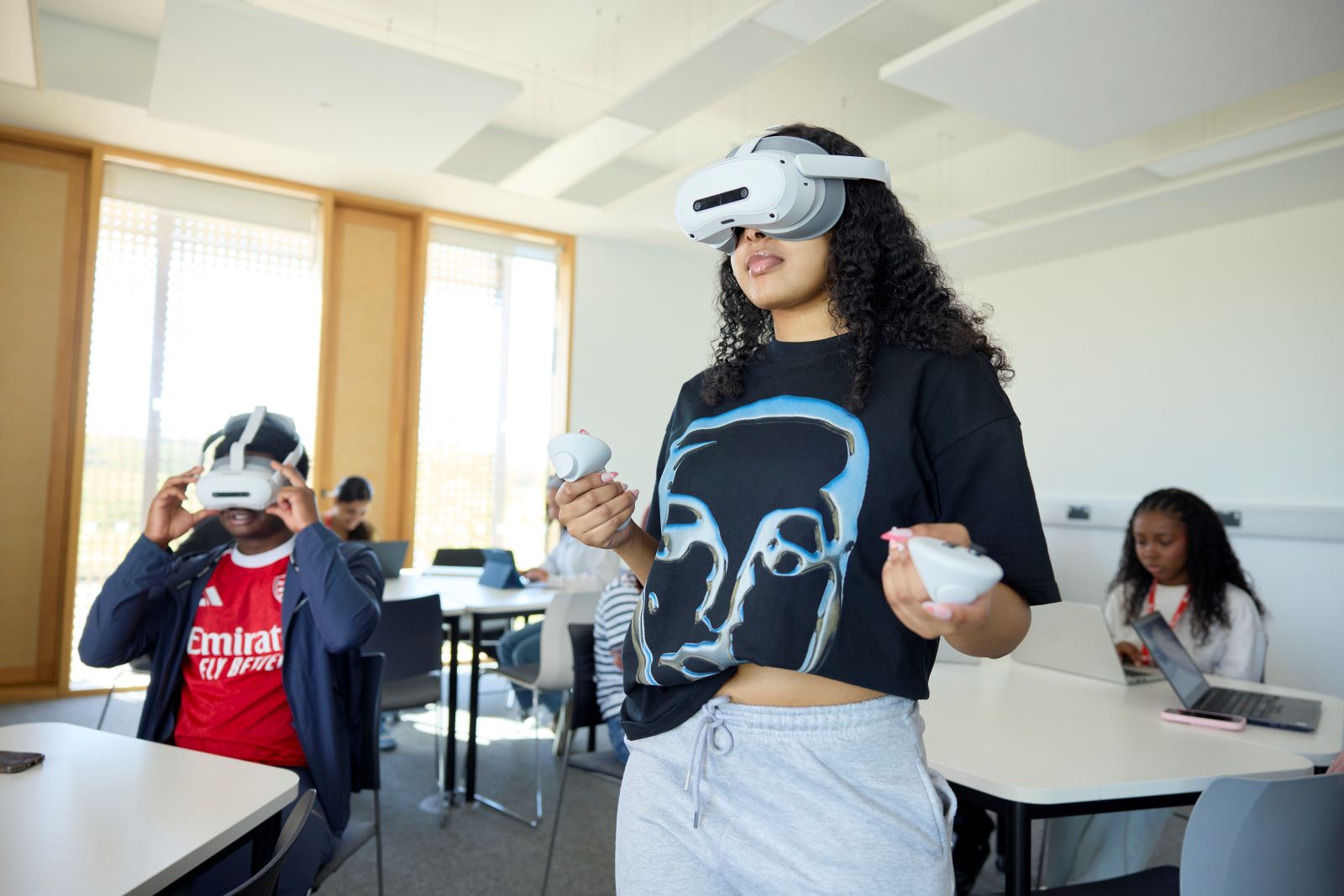 Two students wear virtual reality headsets in a classroom. One is standing and holds a controller in each hand, while the other sits at a table. In the background, other students sits at tables and work on laptops.
