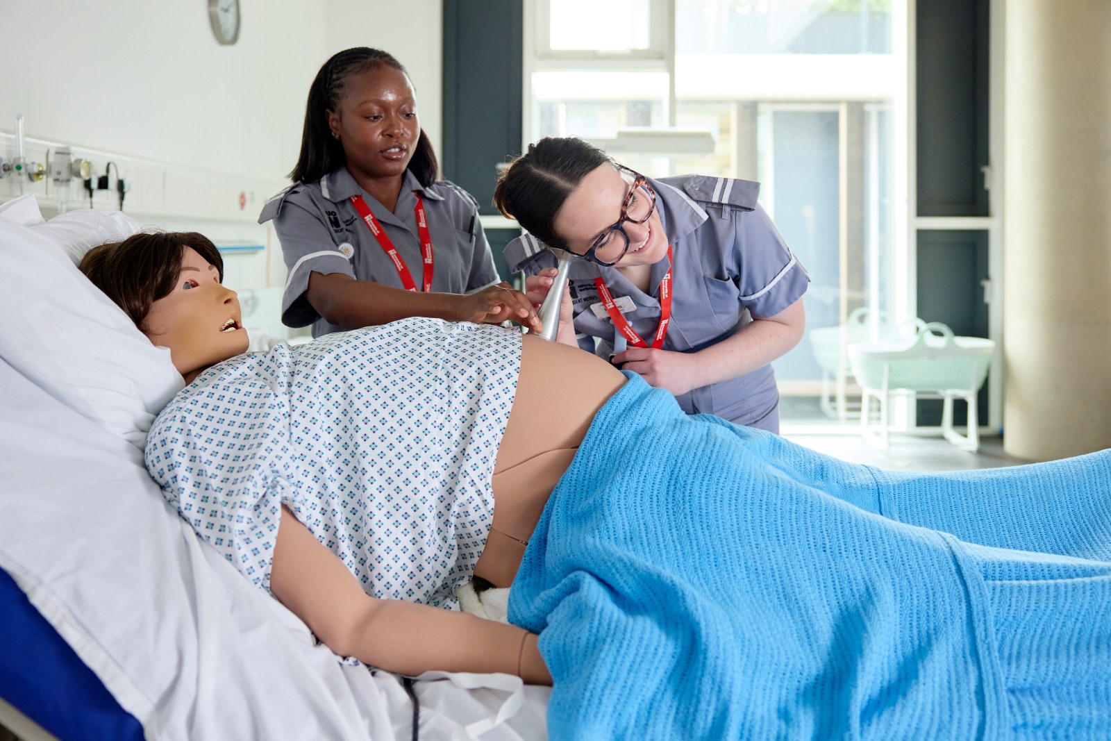 Two healthcare students use a fetal monitoring device on a maternity simulation mannequin in a clinical training room.