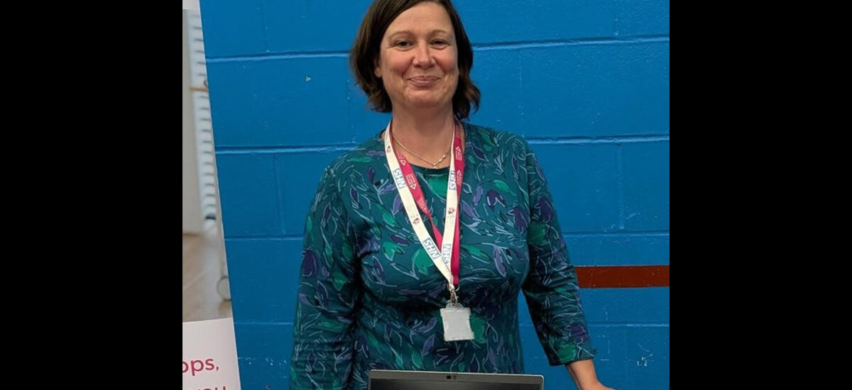 Helen Macmillan, CPD student. She is wearing a dark green and purple patterned dress and is standing in front of a blue wall.