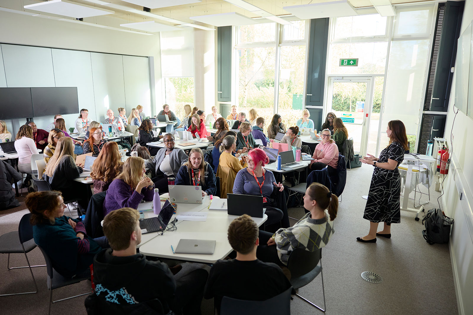 Students seated at round tables in a classroom, actively engaged in a lecture, listening, discussing ideas, and participating in collaborative learning activities while the lecturer leads the session at the front of the room.