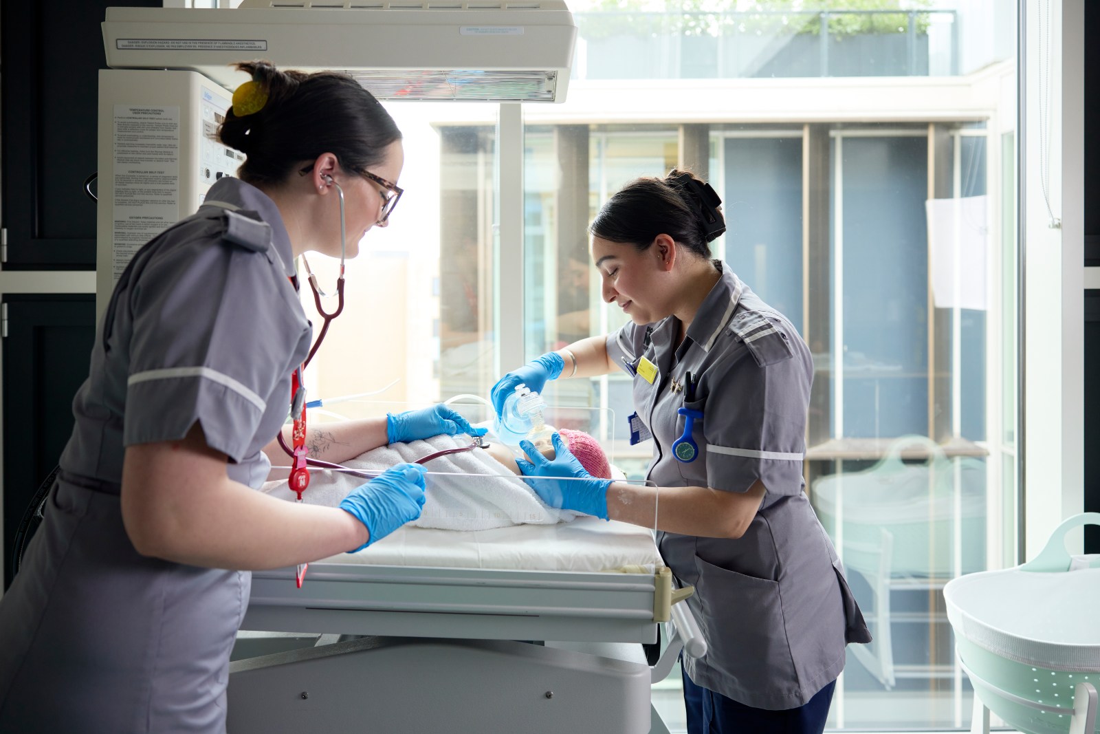 Two healthcare students in clinical uniforms provide newborn care at an incubator, with one using a stethoscope while the other supports the baby's breathing.