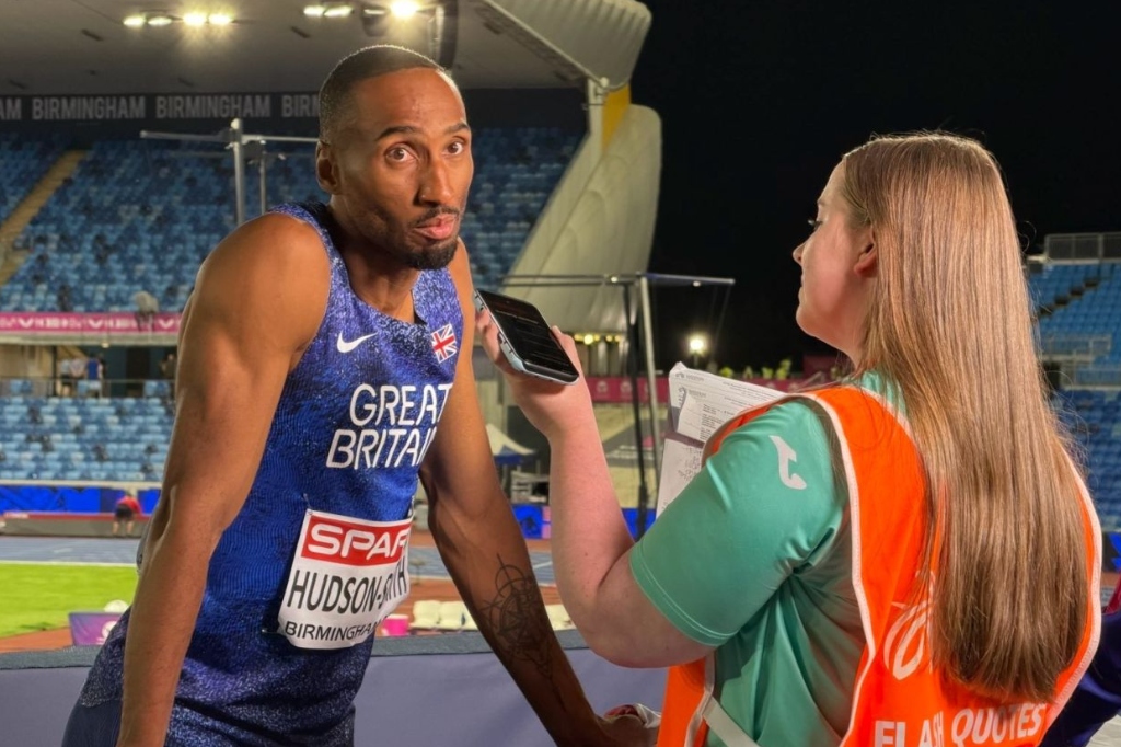 Student in orange bib holds microphone up to athlete in interview.
