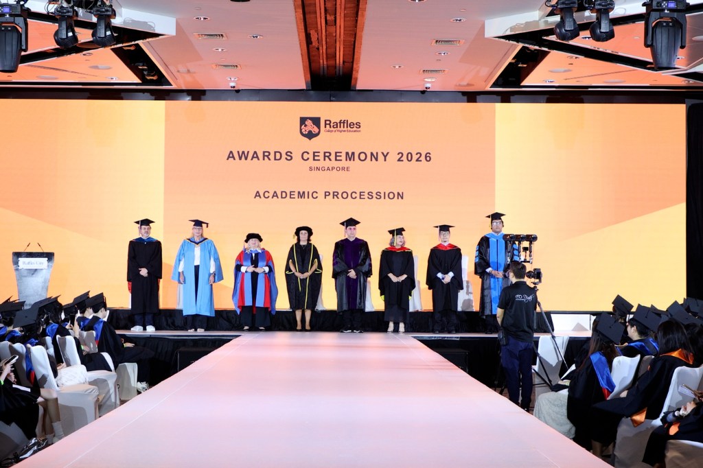 Eight people in graduation gowns stand on a stage. A sign for 'Raffles GRADuation' is displayed behind them, with graduands sitting either side of the stage runway.