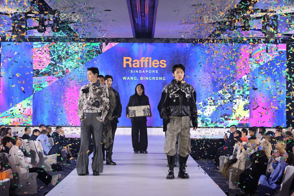Five people stand on a runway stage. Confetti falls around them, and the winning student stands in the centre of the stage holding their certificate and smiling.