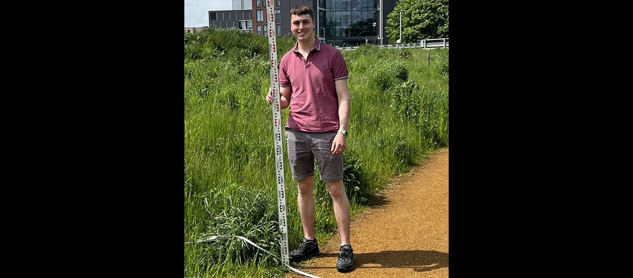 Matthew Vickers, Geography BSc graduate. He is wearing a t-shirt and shorts and is standing on an outside path while holding a measuring tool.