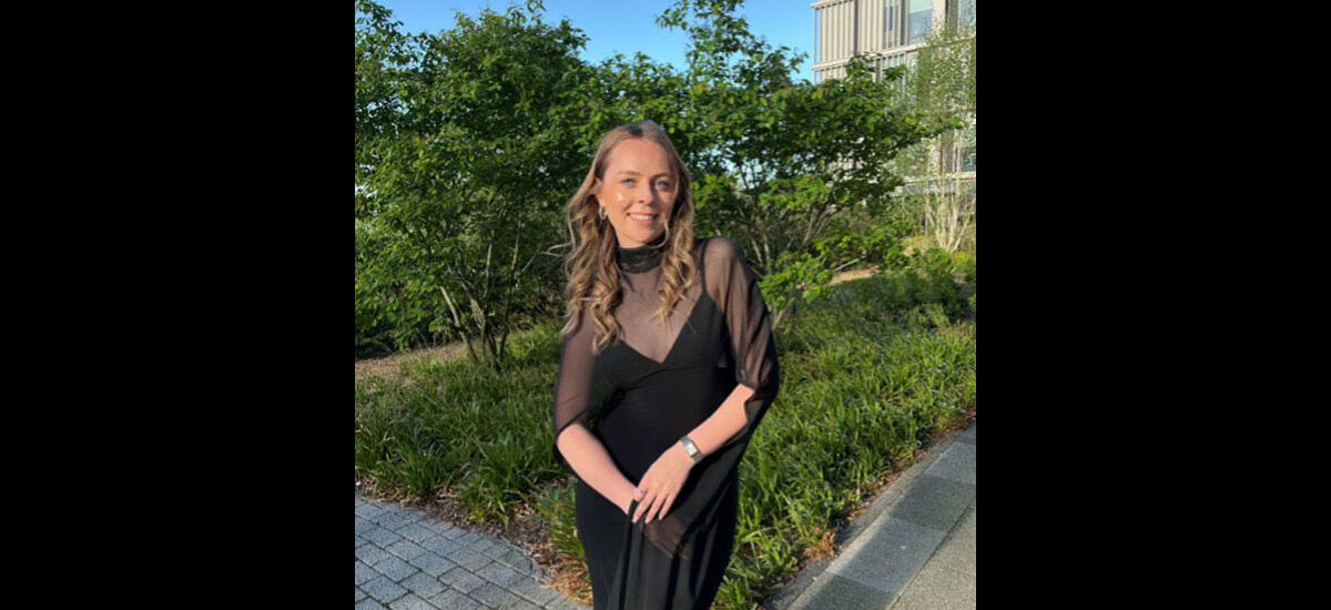 Jana Nietzke, International Marketing Strategy MSc graduate. She is wearing a long black dress and is standing in front of some small trees on Waterside campus.