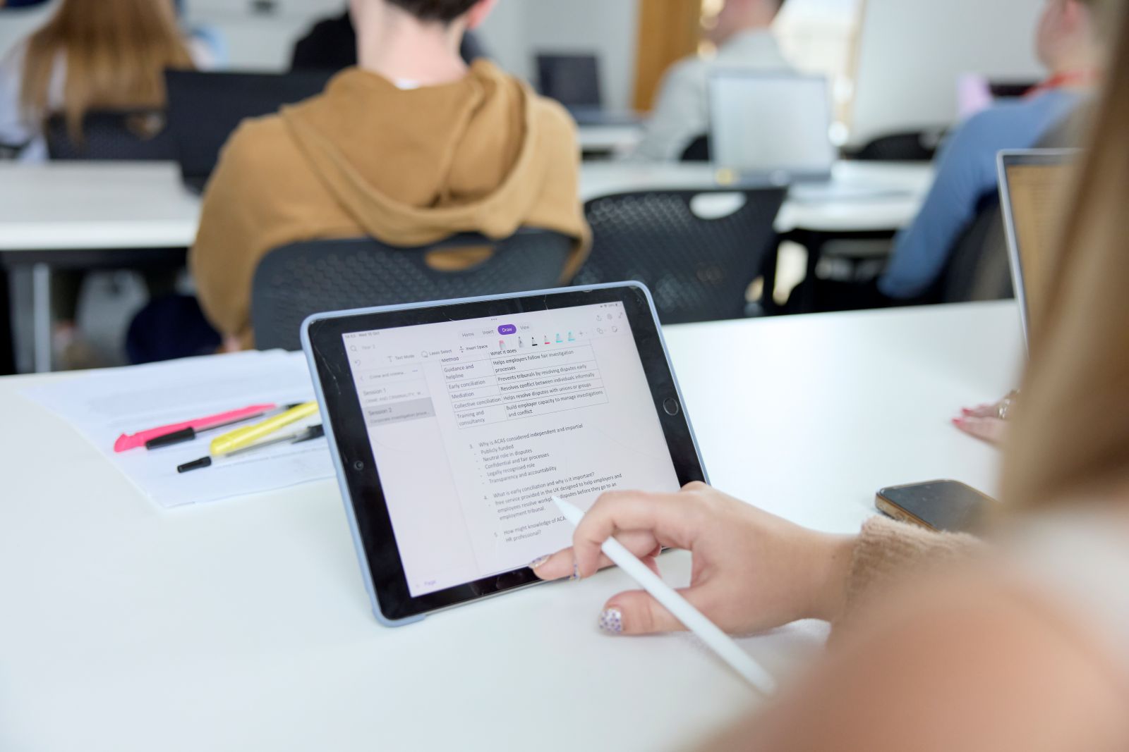 Student uses a tablet in a classroom during a seminar. The student holds a white pen in their hand and is wearing metallic nail polish. In front of them, other students sit at tables.