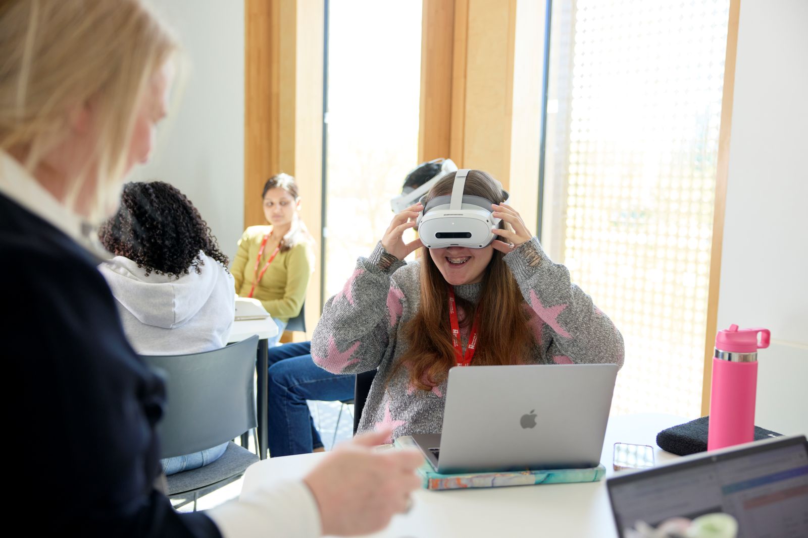 A student wears a virtual reality headset during a seminar. She is seated at a table and is wearing a grey jumper with a pink star pattern. There is an open laptop and a pink water bottle on the table. In the foreground, an academic is standing, while behind her, other students are seated at tables.