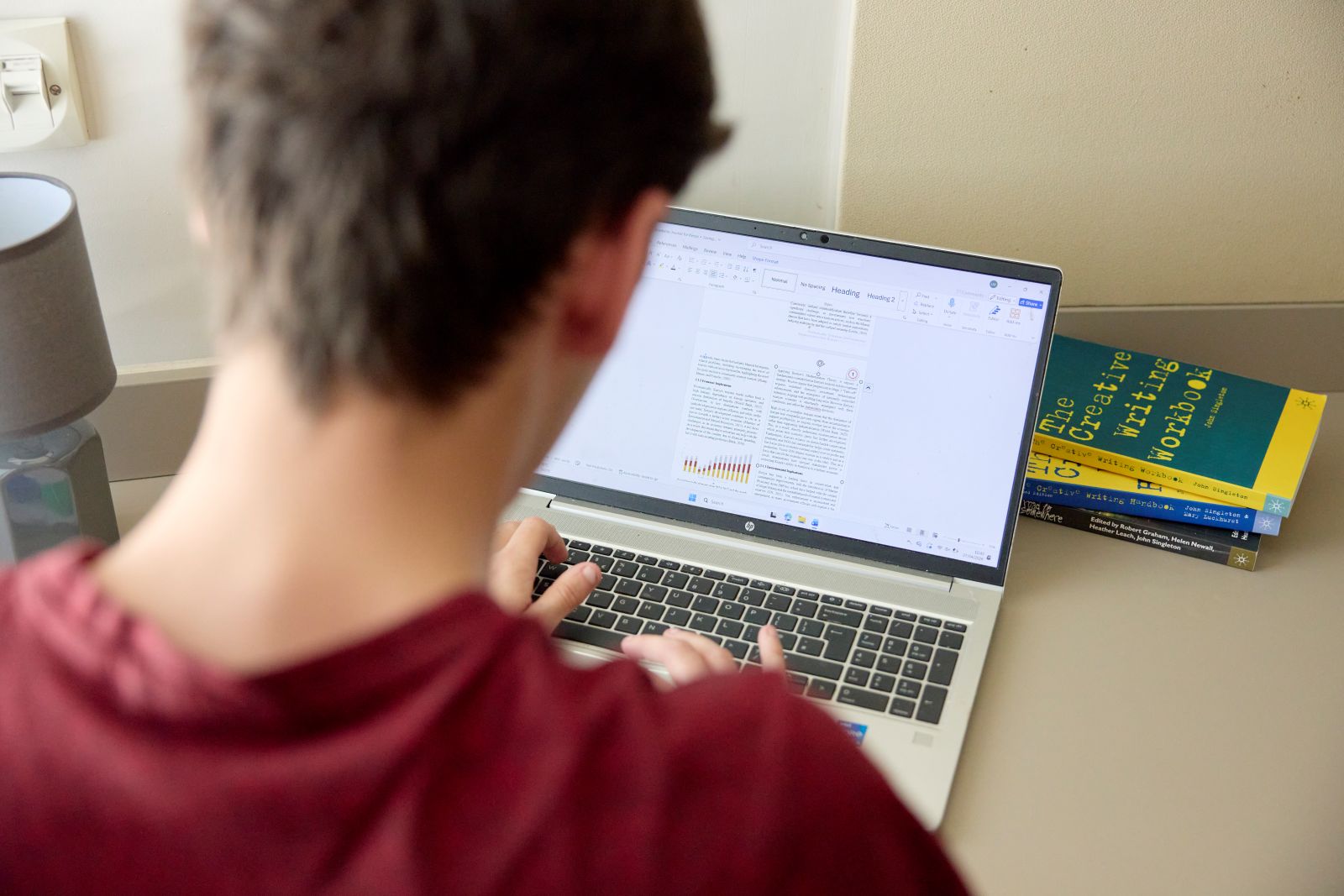 Over-the-shoulder photograph of a student working on a laptop at a desk. The laptop screen shows a document with text and charts being edited. A lamp is positioned on the left side of the desk, and several creative writing books are stacked beside the computer.