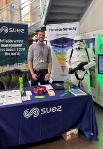 A Suez representative stands at a branded information table at an environmental awareness event in the market restaurant, alongside a stormtrooper character costume. The display includes promotional materials, reusable bottles and a banner explaining the waste hierarchy.