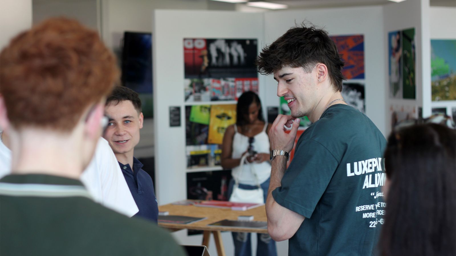 Students and visitors discussing creative work in a studio space, with photography and visual design projects displayed on exhibition panels in the background.