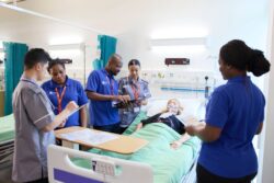 Five healthcare workers stand around a hospital bed, discussing notes, while a patient wearing an oxygen mask lies in the bed.