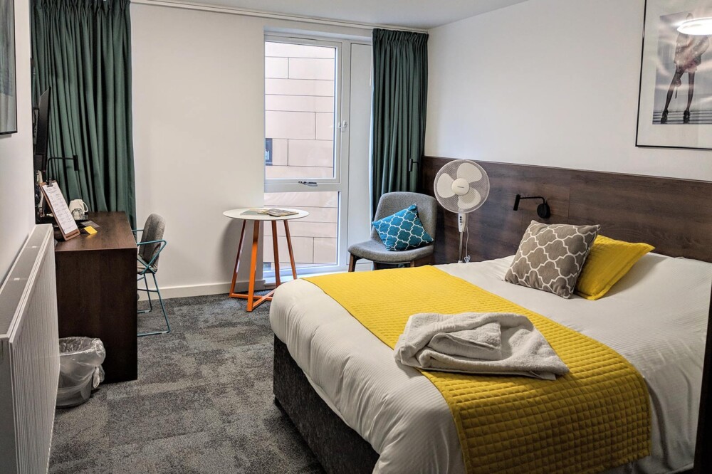 View of a bedroom at Sunley Hotel, showing a double bed with a white and yellow cover and a towel, a standing fan, a desk with a chair, and a small table with another chair.