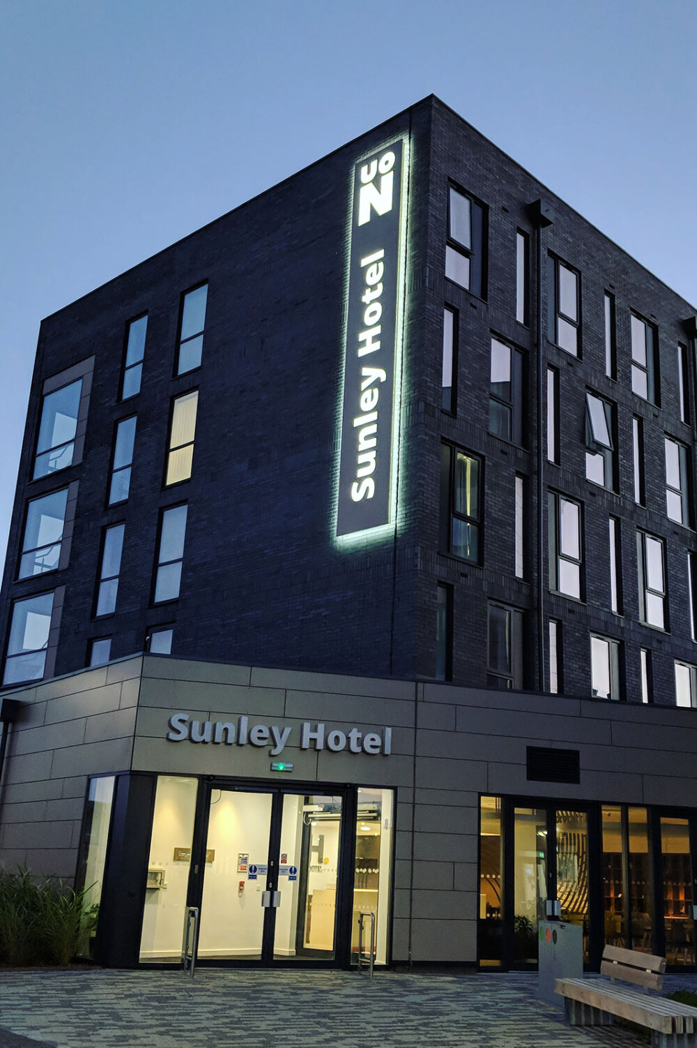 External view of Sunley Hotel at night, so the light is on the vertical sign on the building and there is light coming from the reception in the windows