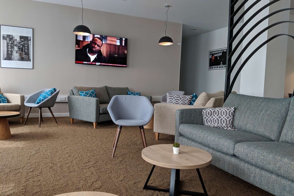 View of the Sunley hotel foyer, showing three different types of chair: sofas in cream and grey, and an individual chair in light grey. There are a couple of low coffee tables between and a TV on the wall.