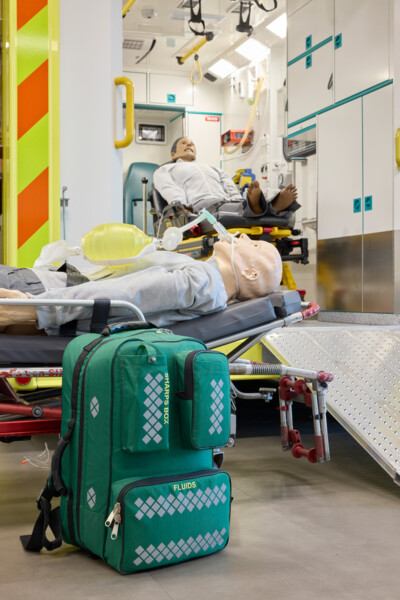 Photo of two mannequins in the Ambulance simulator. Both are on stretchers, one inside the ambulance and one outside. There is a backpack next to one, with the fluids and sharps labelled.